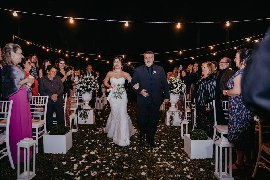 A bride in a strapless white wedding gown holding a bouquet of white and blue flowers is walking down the aisle, escorted by a man in a dark suit. The aisle is adorned with white lanterns and small ma