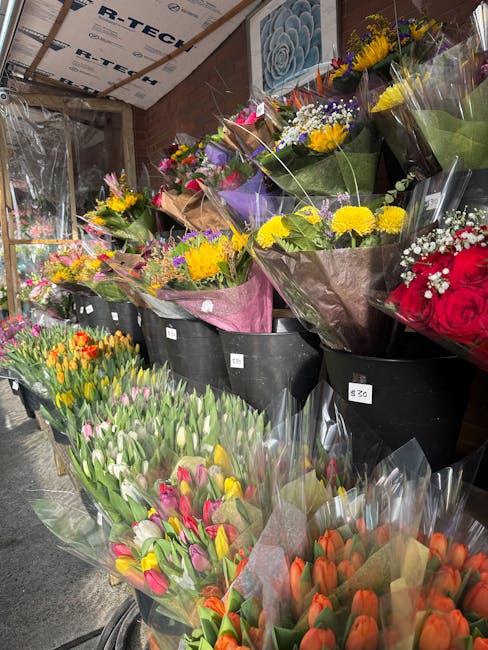A diverse selection of fresh flower bouquets arranged in black buckets and wrapped in colorful paper and cellophane, featuring yellow, purple, red, pink, and orange blooms such as roses, tulips, daffo