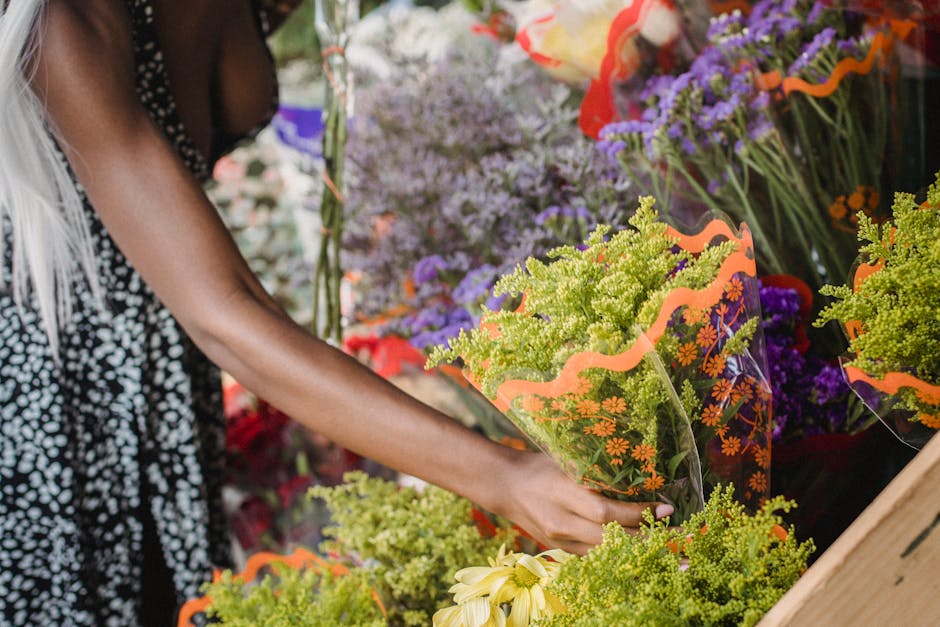 A person selecting a vibrant bouquet of fresh mixed flowers at a florist shop, with various other flower arrangements displayed in the background. The bouquet in their hand features clusters of yellow