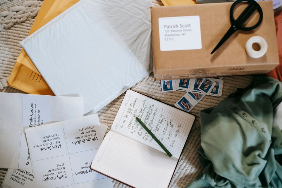 A flat lay arrangement on a textured beige surface showcasing various mailing and packaging materials, including white and yellow envelopes, a brown cardboard box labeled with a recipient address, a p