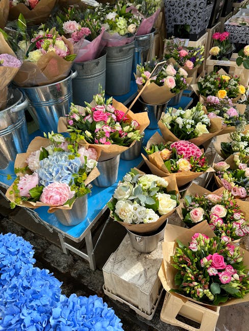 A variety of fresh floral bouquets arranged in metal and ceramic vases at a flower market, featuring pastel pink and white roses, blue hydrangeas, and green foliage. The bouquets are wrapped in brown 